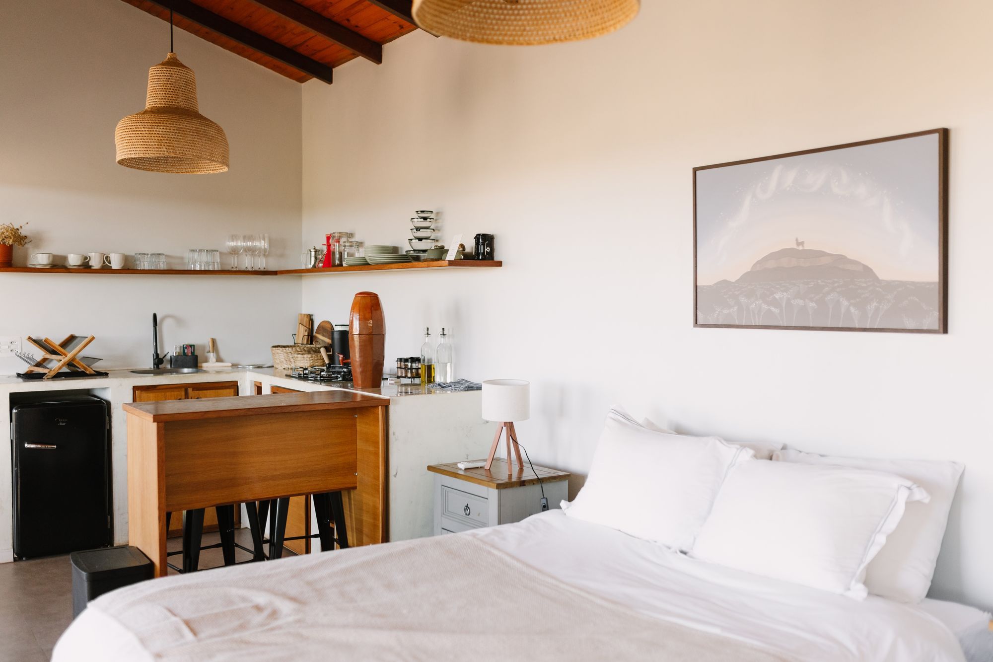 Cama queen com roupa de cama branca e, ao fundo, a cozinha do Chalé Deluxe c/ Hidromassagem com frigobar retrô e bancada de madeira