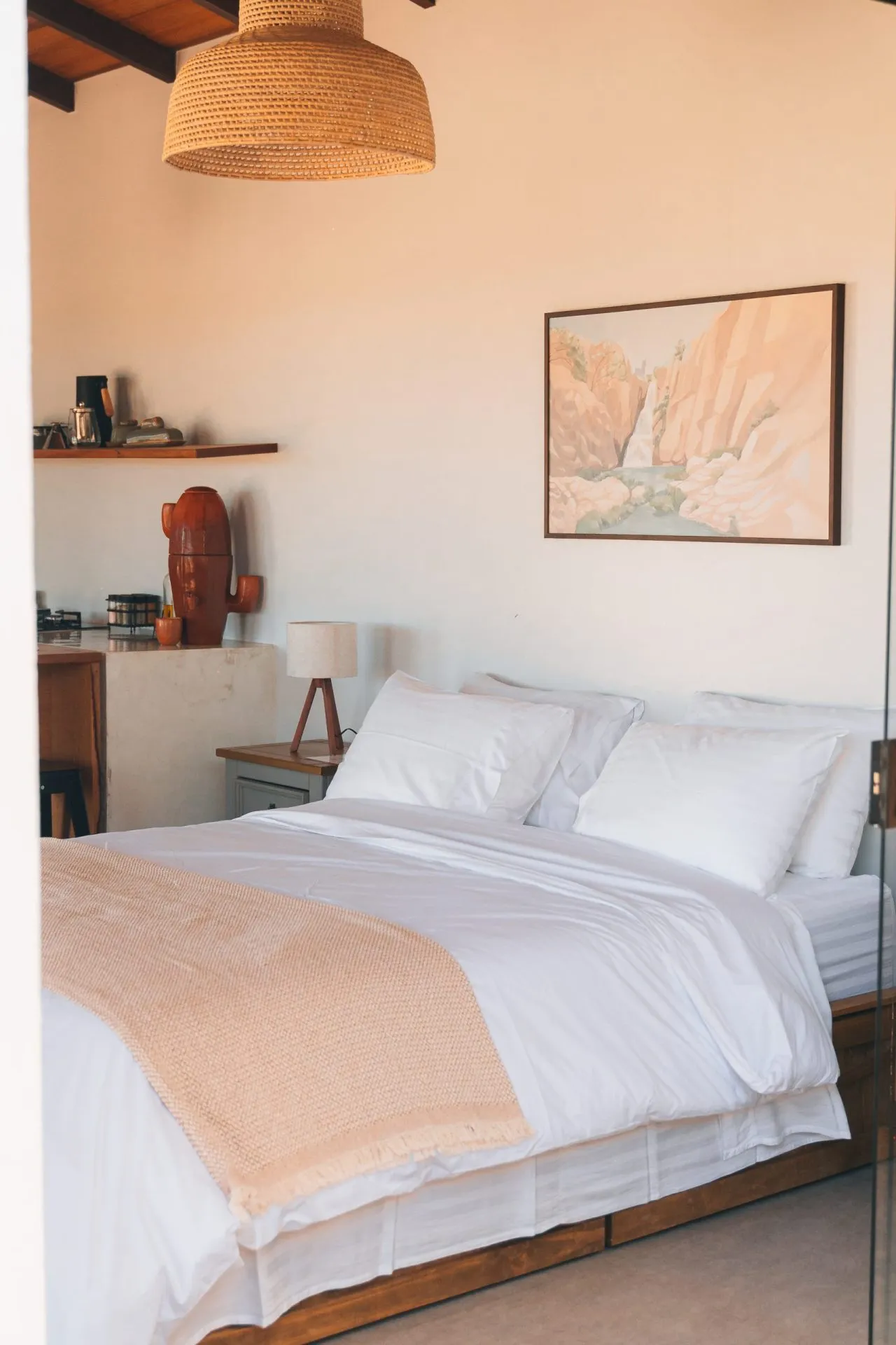 Quarto do chalé com cama king de lençóis brancos, pendente de rattan, quadro de paisagem e cabeceira de madeira