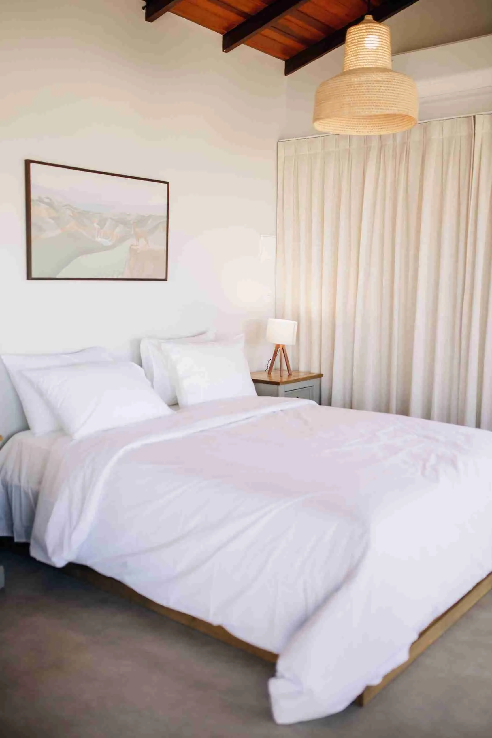 Cama com lençóis brancos e luminária de tripé, cortinas de linho e pendente de palha no teto de madeira aparente