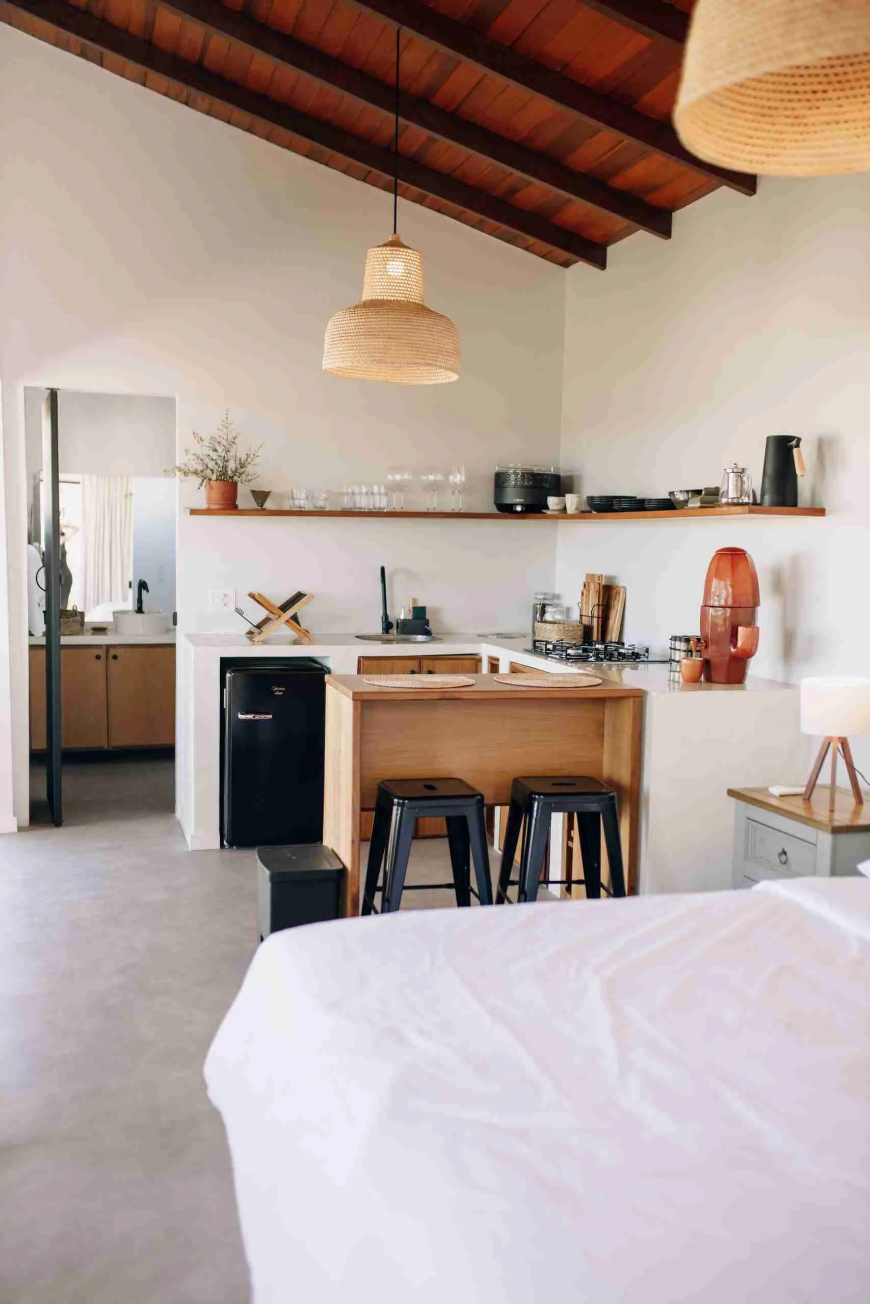 Interior completo do chalé com cama, cozinha com ilha de madeira, banquetas pretas, geladeira retrô e pendentes de rattan