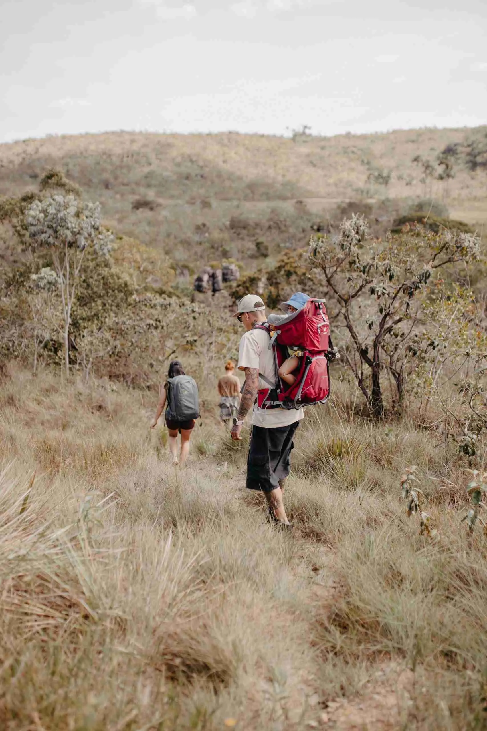 Família fazendo trilha pelo Cerrado, pai carregando bebê na mochila canguru vermelha