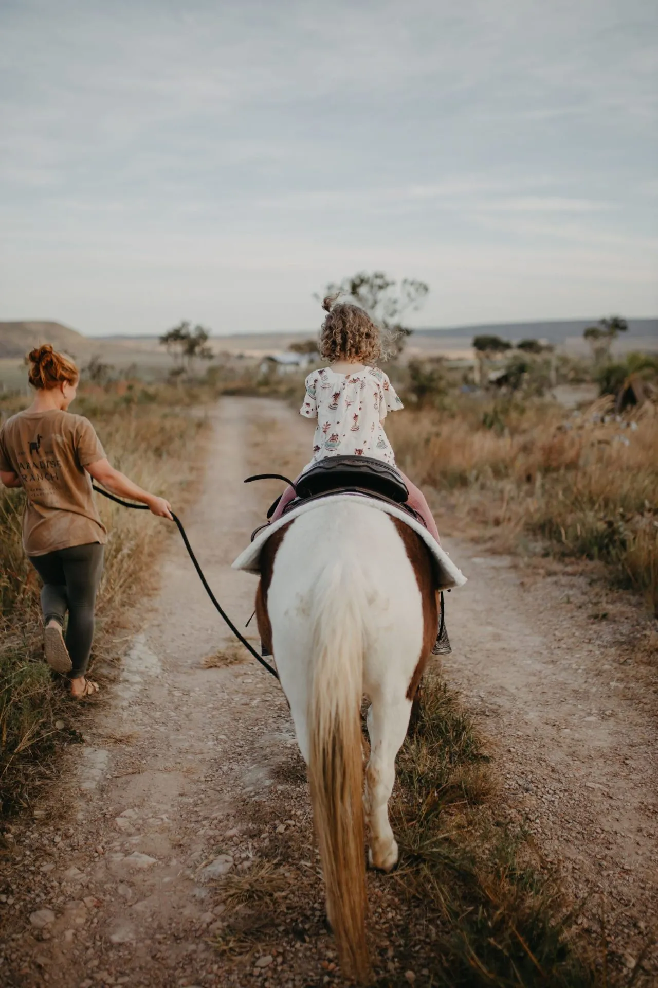 Criança passeando de pônei guiado por funcionária do rancho em trilha de terra no Cerrado
